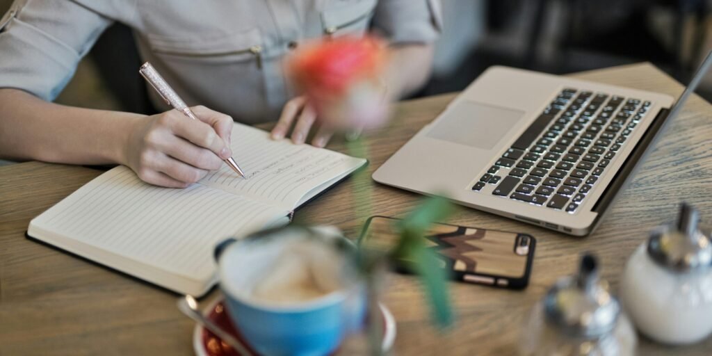 Woman writing in a notebook with a laptop and coffee cup on a desk. Ideal for workspace inspiration.