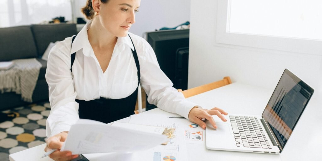 Woman at desk reviewing business reports on laptop, holding papers in a modern office setting.