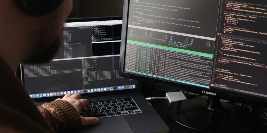 Focused programmer coding at dual monitors with headphones, using a laptop and desktop setup for efficient software development.